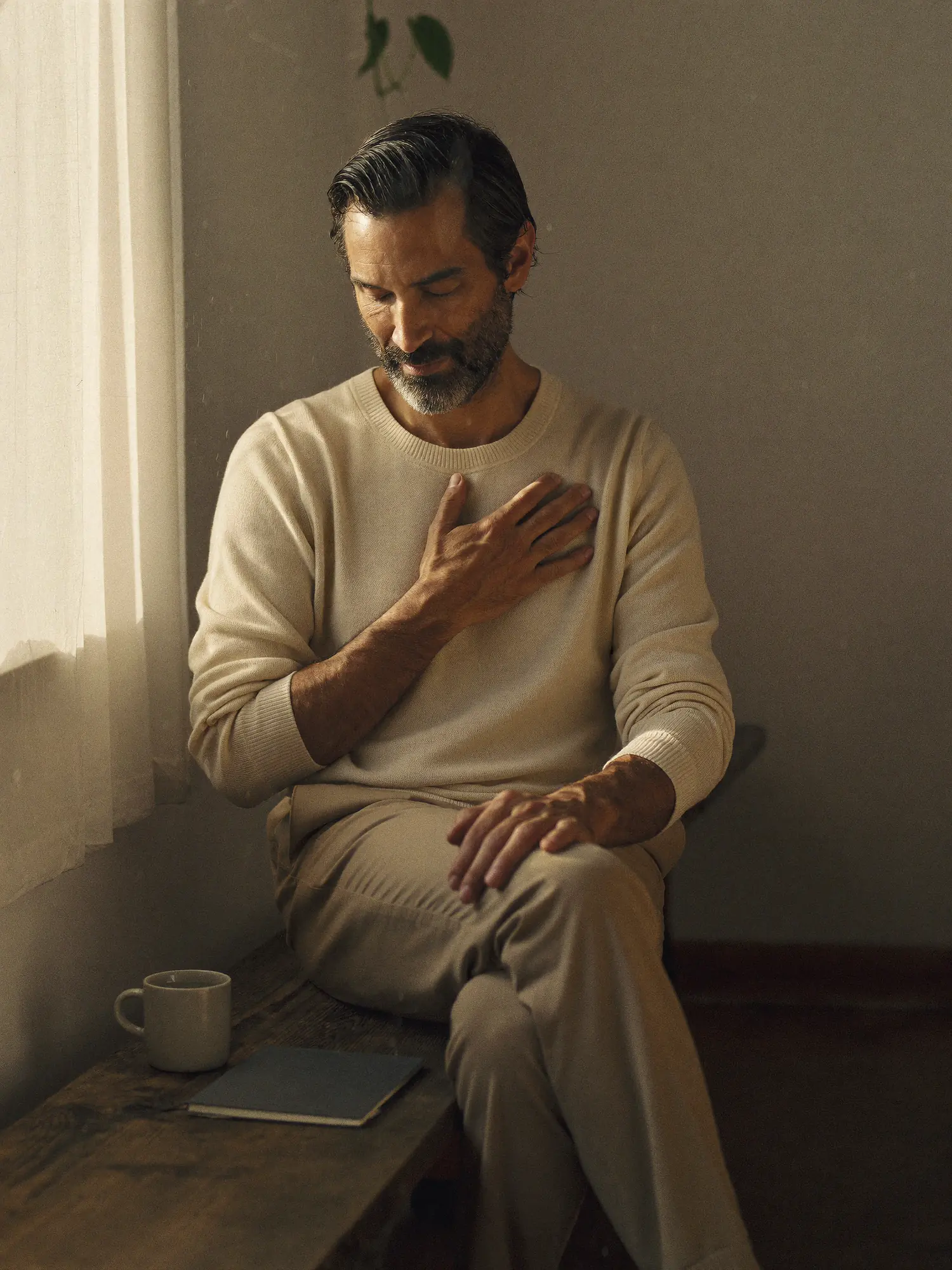 A man sits on a wooden bench in a sunlit room, eyes closed, one hand resting on his chest