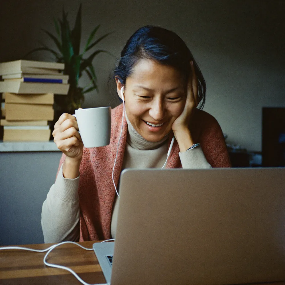 A woman laughs as she joins an online class from her kitchen table, mug in hand