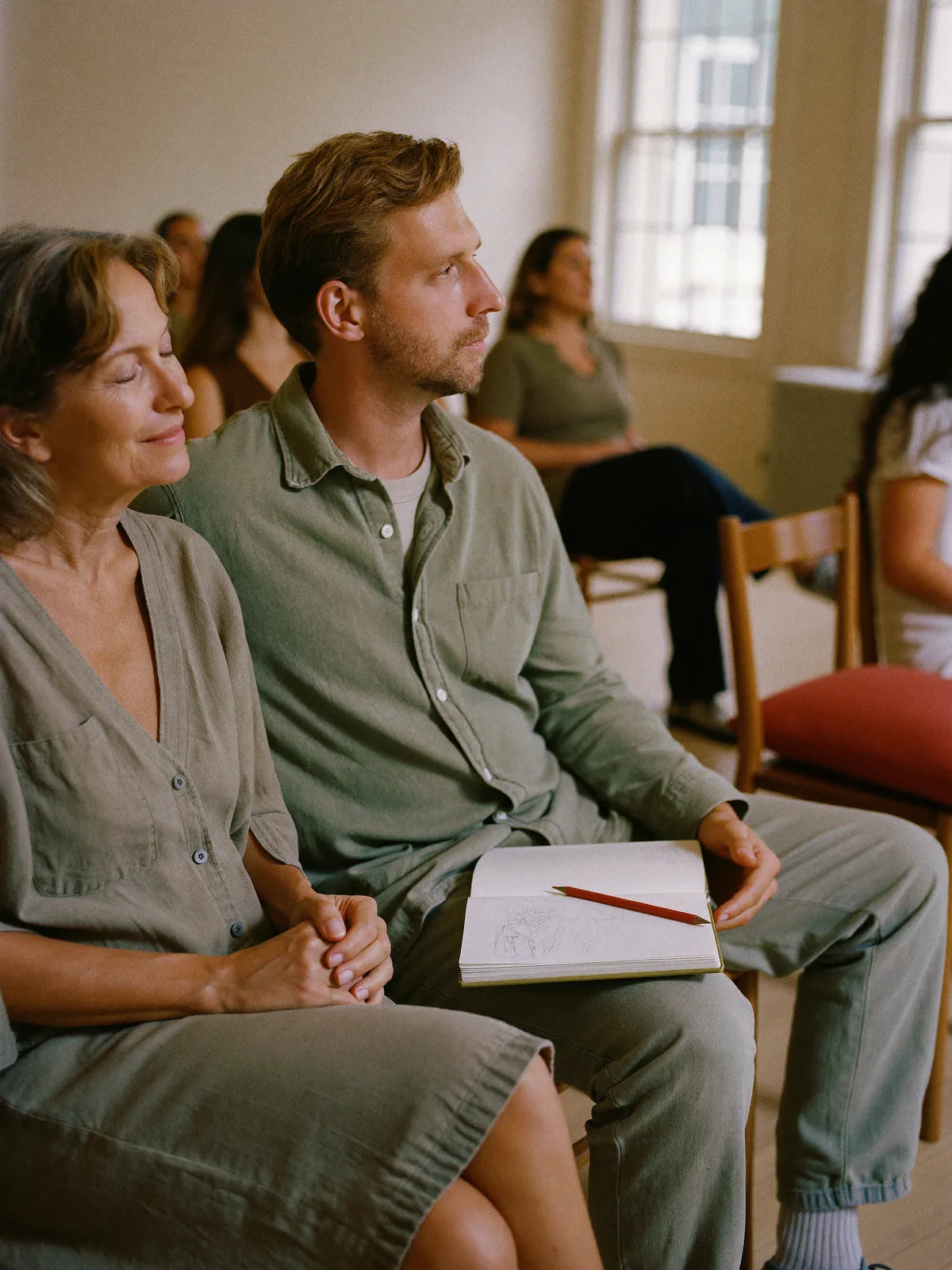Two adults sit side by side in a training room, one with eyes closed, the other holding a notebook